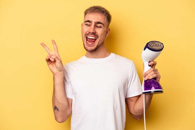 Young caucasian man holding an iron isolated on yellow background joyful and carefree showing a peace symbol with fingers.