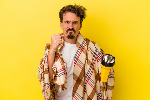 Young caucasian man holding lantern isolated on yellow background showing fist to camera, aggressive facial expression.