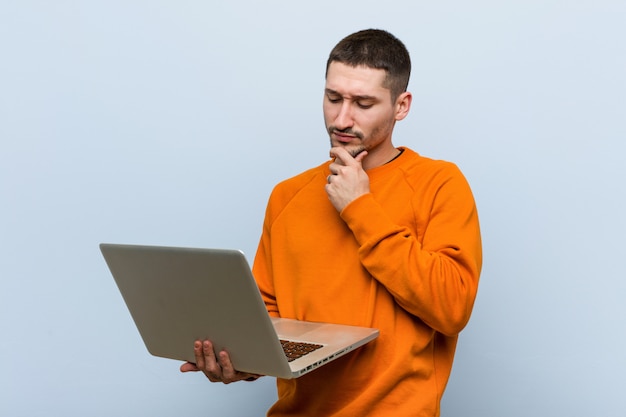 Young caucasian man holding a laptop looking sideways with doubtful and skeptical expression.