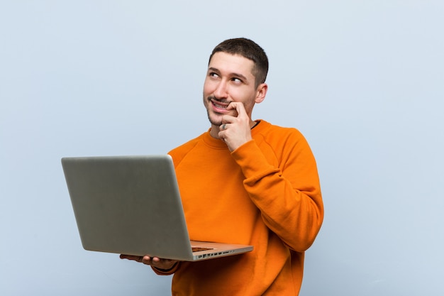 Young caucasian man holding a laptop relaxed thinking about something looking at a copy space.