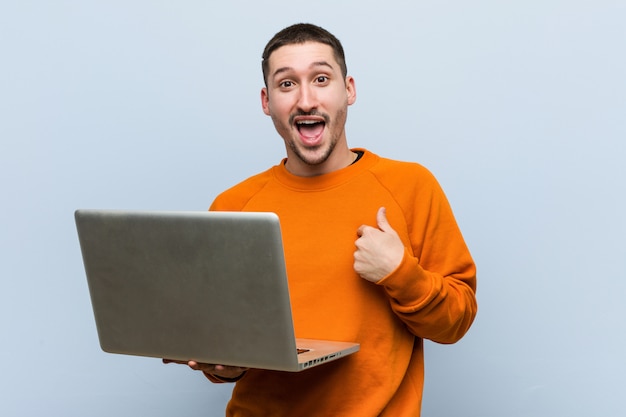 Il giovane uomo caucasico che tiene un computer portatile sorpreso indicando se stesso, sorridendo ampiamente.
