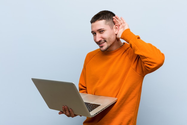 Young caucasian man holding a laptop trying to listening a gossip.