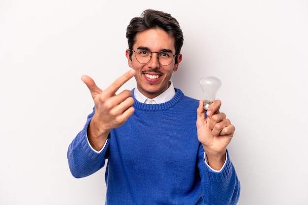 Young caucasian man holding a lightbulb isolated on white background pointing with finger at you as if inviting come closer