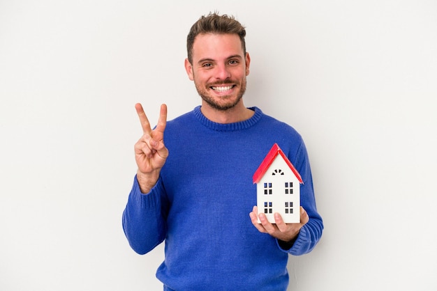 Young caucasian man holding a little house isolated on white background joyful and carefree showing a peace symbol with fingers