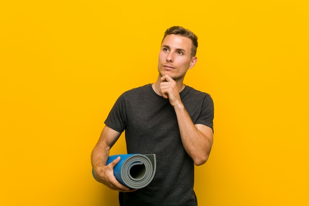 Young caucasian man holding a mat looking sideways with doubtful and skeptical expression