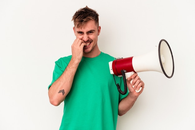 Young caucasian man holding megaphone isolated on white background biting fingernails, nervous and very anxious.