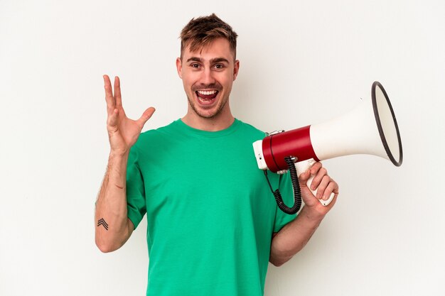 Young caucasian man holding megaphone isolated on white background receiving a pleasant surprise, excited and raising hands.