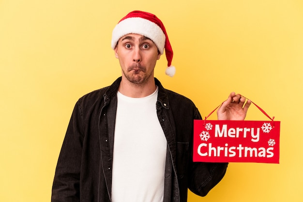 Young caucasian man holding a Merry Christmas placard isolated on yellow background shrugs shoulders and open eyes confused.