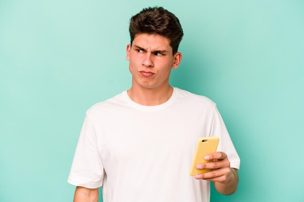 Young caucasian man holding mobile phone isolated on blue background confused feels doubtful and unsure