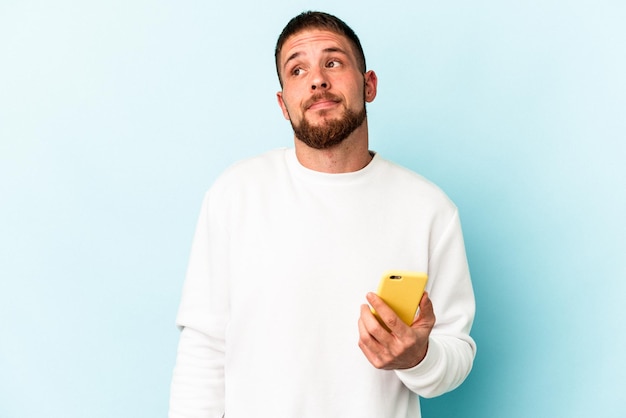 Young caucasian man holding mobile phone isolated on blue background dreaming of achieving goals and purposes