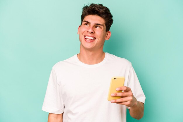 Young caucasian man holding mobile phone isolated on blue background dreaming of achieving goals and purposes