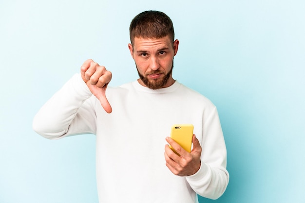 Young caucasian man holding mobile phone isolated on blue background showing a dislike gesture, thumbs down. Disagreement concept.