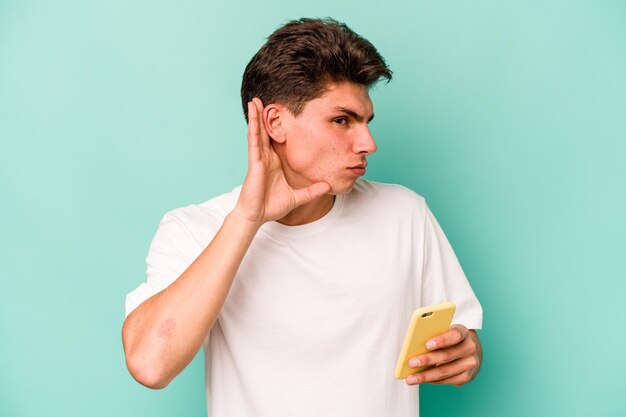 Young caucasian man holding mobile phone isolated on blue background trying to listening a gossip