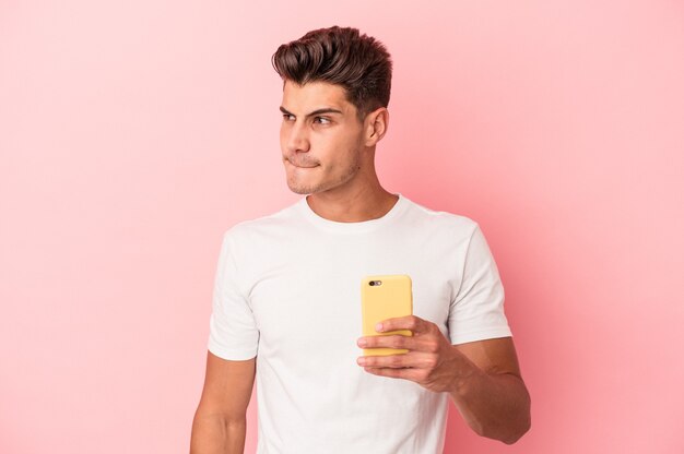 Il giovane uomo caucasico che tiene un telefono cellulare isolato su sfondo rosa confuso, si sente dubbioso e insicuro.