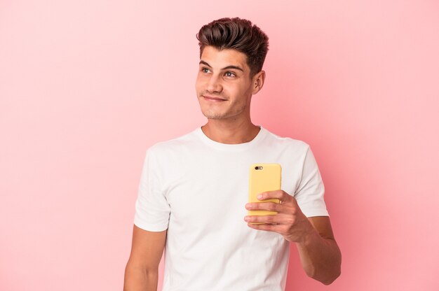 Young caucasian man holding a mobile phone isolated on pink background dreaming of achieving goals and purposes