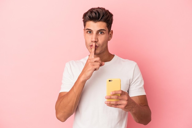 Young caucasian man holding a mobile phone isolated on pink background keeping a secret or asking for silence.