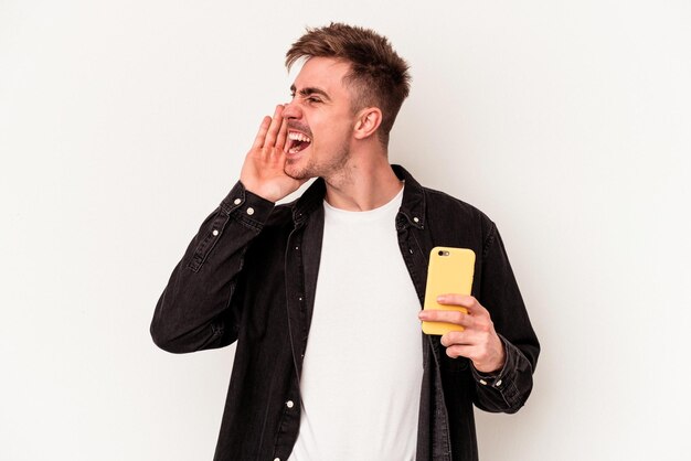 Молодой кавказский человек, держащий мобильный телефон, изолированные на белом фоне, кричит и держит ладонь возле открытого рта.