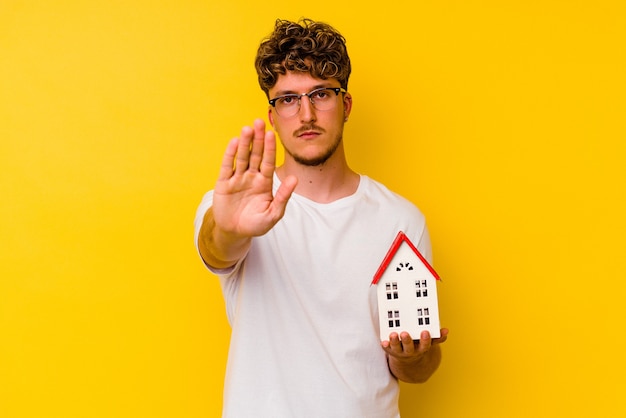 Young caucasian man holding a model house isolated on yellow background standing with outstretched hand showing stop sign, preventing you.