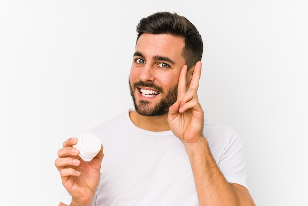 Young caucasian man holding a moisturizer showing victory sign and smiling broadly.