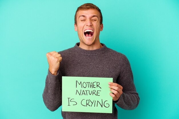 Young caucasian man holding a Mother Nature is crying placard isolated on blue cheering carefree and excited. Victory concept.