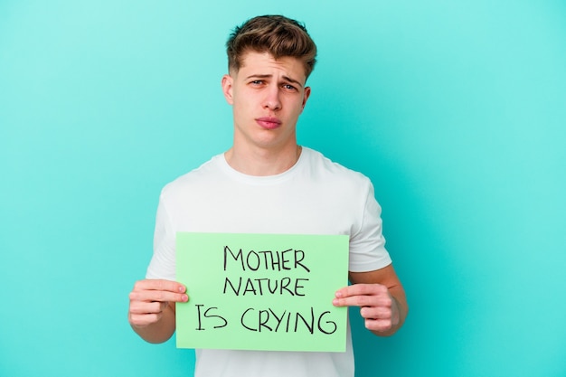 Young caucasian man holding a Mother Nature is crying placard isolated on blue wall