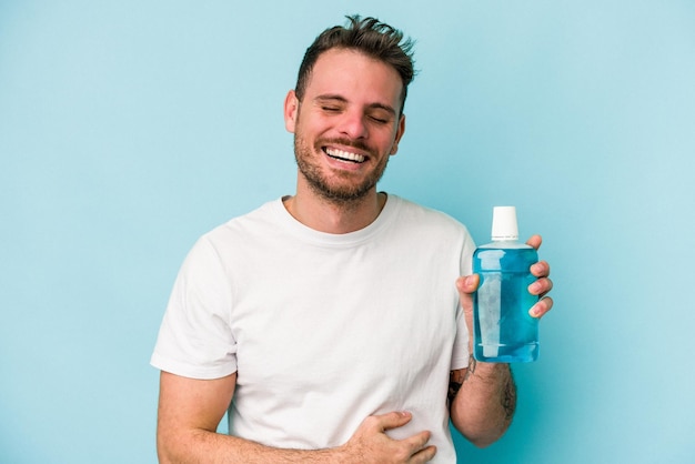 Young caucasian man holding mouthwash isolated on blue background laughing and having fun