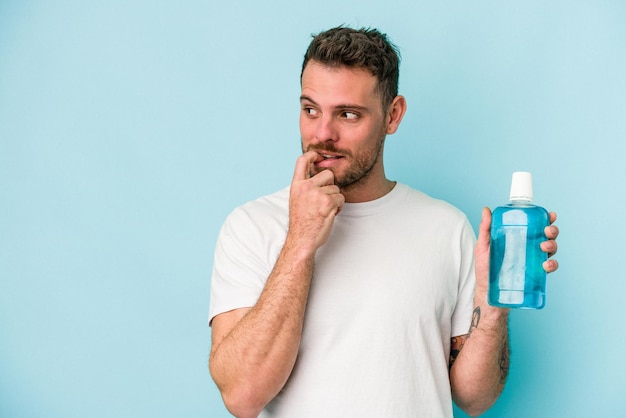 Young caucasian man holding mouthwash isolated on blue background relaxed thinking about something looking at a copy space.