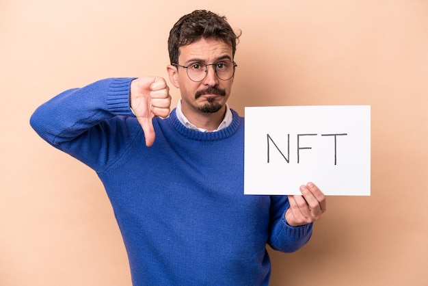 Young caucasian man holding a NFT placard isolated on beige background showing a dislike gesture, thumbs down. Disagreement concept.