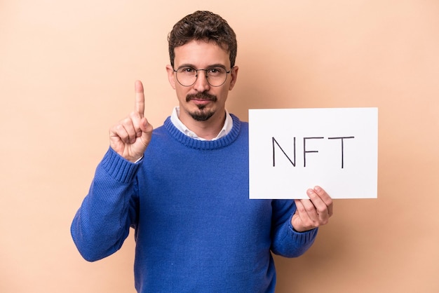 Young caucasian man holding a NFT placard isolated on beige background showing number one with finger.