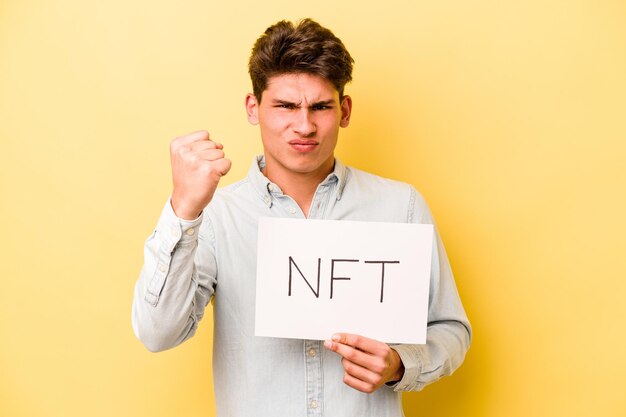 Young caucasian man holding NFT placard isolated on yellow background showing fist to camera aggressive facial expression