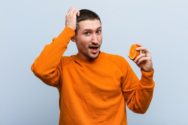 Young caucasian man holding an orange being shocked, she has remembered important meeting.