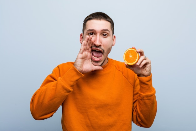 Young caucasian man holding an orange shouting excited to front.