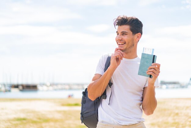 Young caucasian man holding a passport at outdoors thinking an idea and looking side