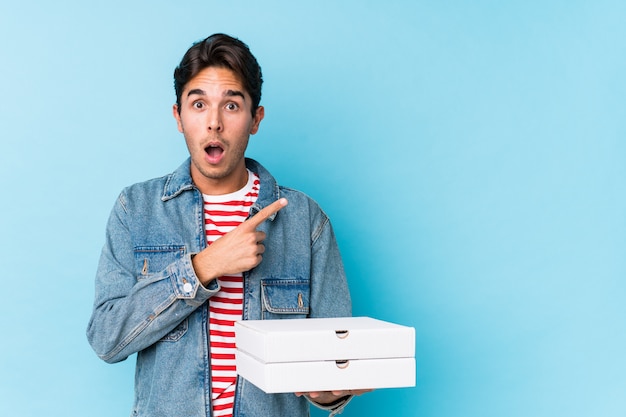 Young caucasian man holding pizzas isolated pointing to the side