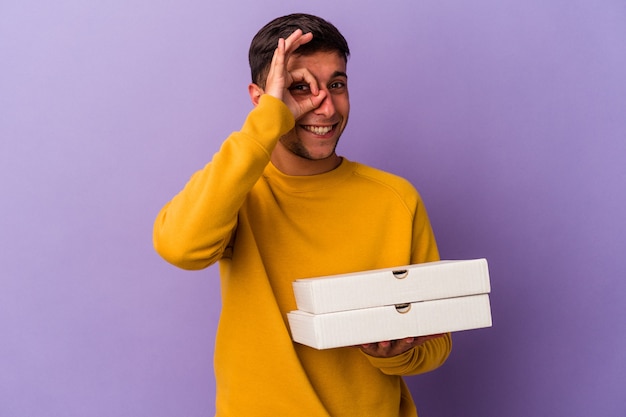 Young caucasian man holding pizzas isolated on purple background excited keeping ok gesture on eye