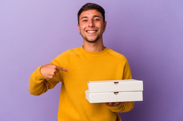 Young caucasian man holding pizzas isolated on purple background  person pointing by hand to a shirt copy space, proud and confident