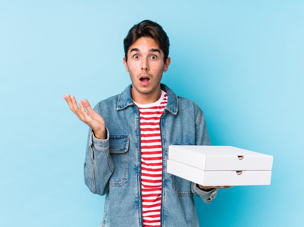 Young caucasian man holding pizzas isolated surprised and shocked.