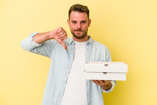 嫌いなジェスチャーを示す黄色の背景に分離されたピザを持っている若い白人男性は、親指を下に向けます。不一致の概念。