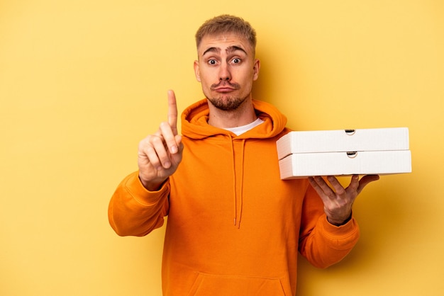 Молодой кавказский мужчина держит пиццу на желтом фоне, показывая пальцем номер один.