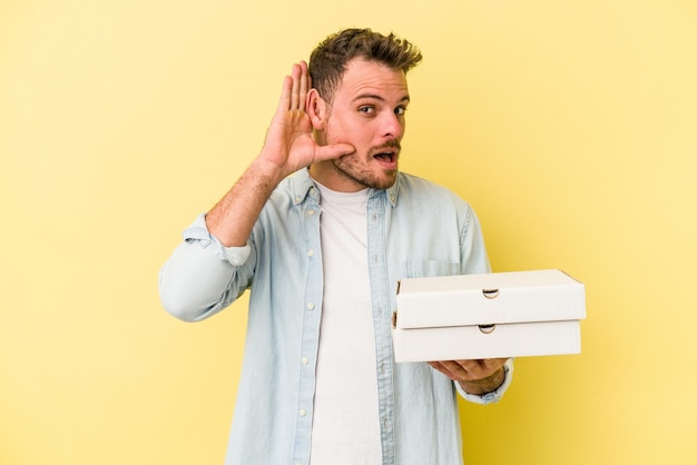 Young caucasian man holding a pizzas isolated on yellow background trying to listening a gossip.