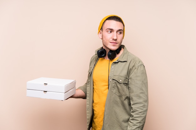 Young caucasian man holding pizzas looks aside smiling, cheerful and pleasant.