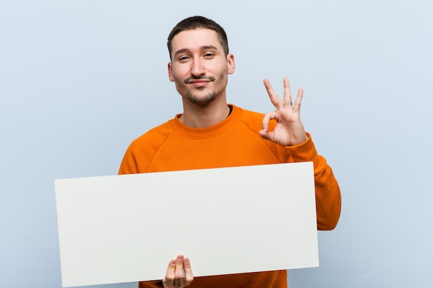 Young caucasian man holding a placard cheerful and confident showing ok gesture.