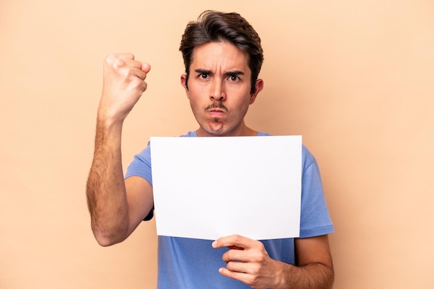 Young caucasian man holding a placard isolated on beige background showing fist to camera, aggressive facial expression.