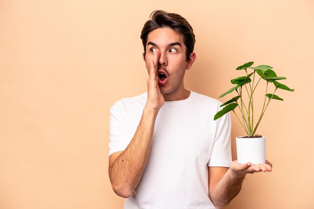 Young caucasian man holding a plant isolated on beige background is saying a secret hot braking news and looking aside