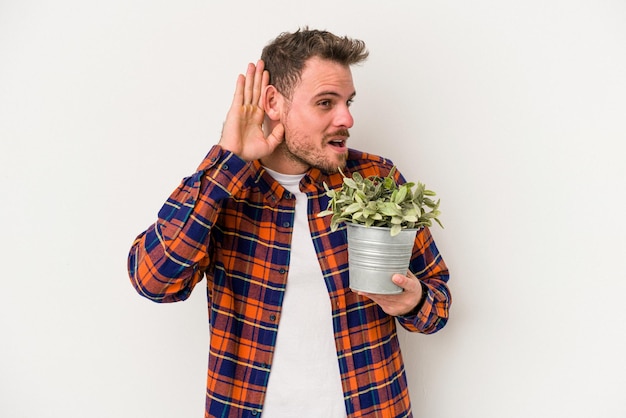 Young caucasian man holding a plant isolated on white background trying to listening a gossip.