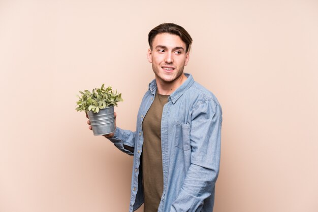 Young caucasian man holding a plant looks aside smiling, cheerful and pleasant.