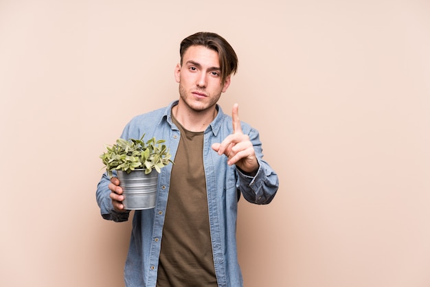 Young caucasian man holding a plant showing number one with finger.