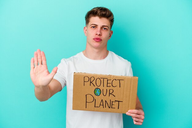 Il giovane uomo caucasico che tiene un protegge il nostro cartello del pianeta isolato su priorità bassa blu
