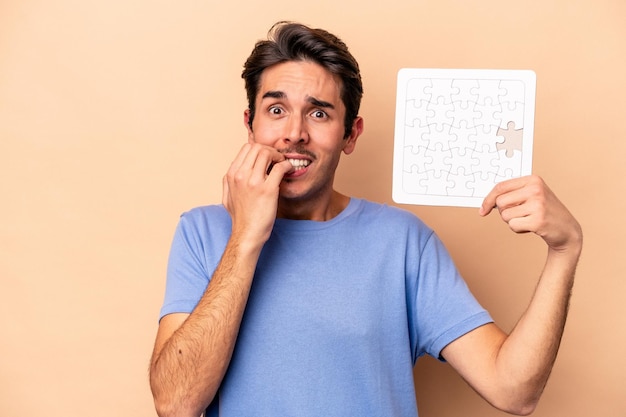 Young caucasian man holding a puzzle isolated on beige background biting fingernails nervous and very anxious