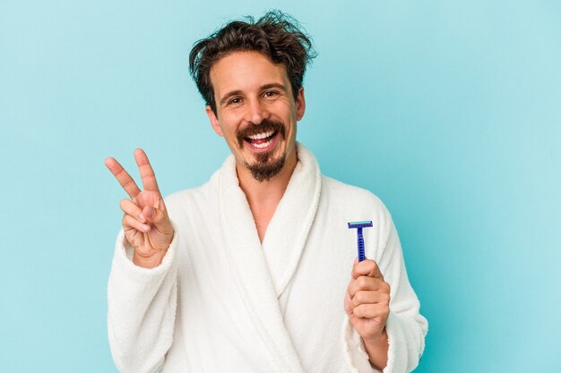 Young caucasian man holding a razor blade isolated on blue background joyful and carefree showing a peace symbol with fingers.